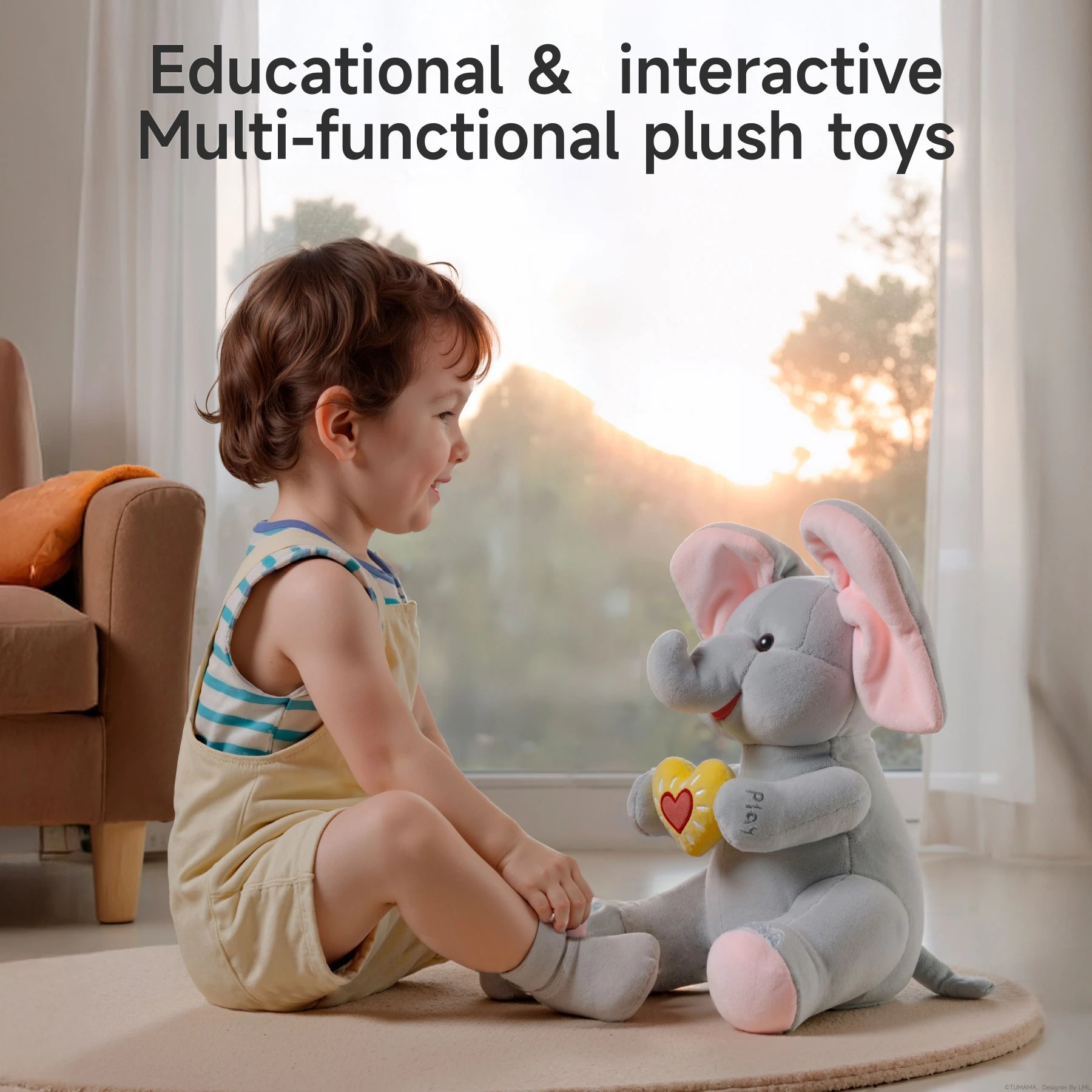 A smiling toddler sitting on the floor facing an interactive elephant plush toy. The elephant has large pink ears and holds a yellow heart with a red center. Text above reads: 'Educational & interactive multi-functional plush toys,' highlighting its learning and entertainment value.