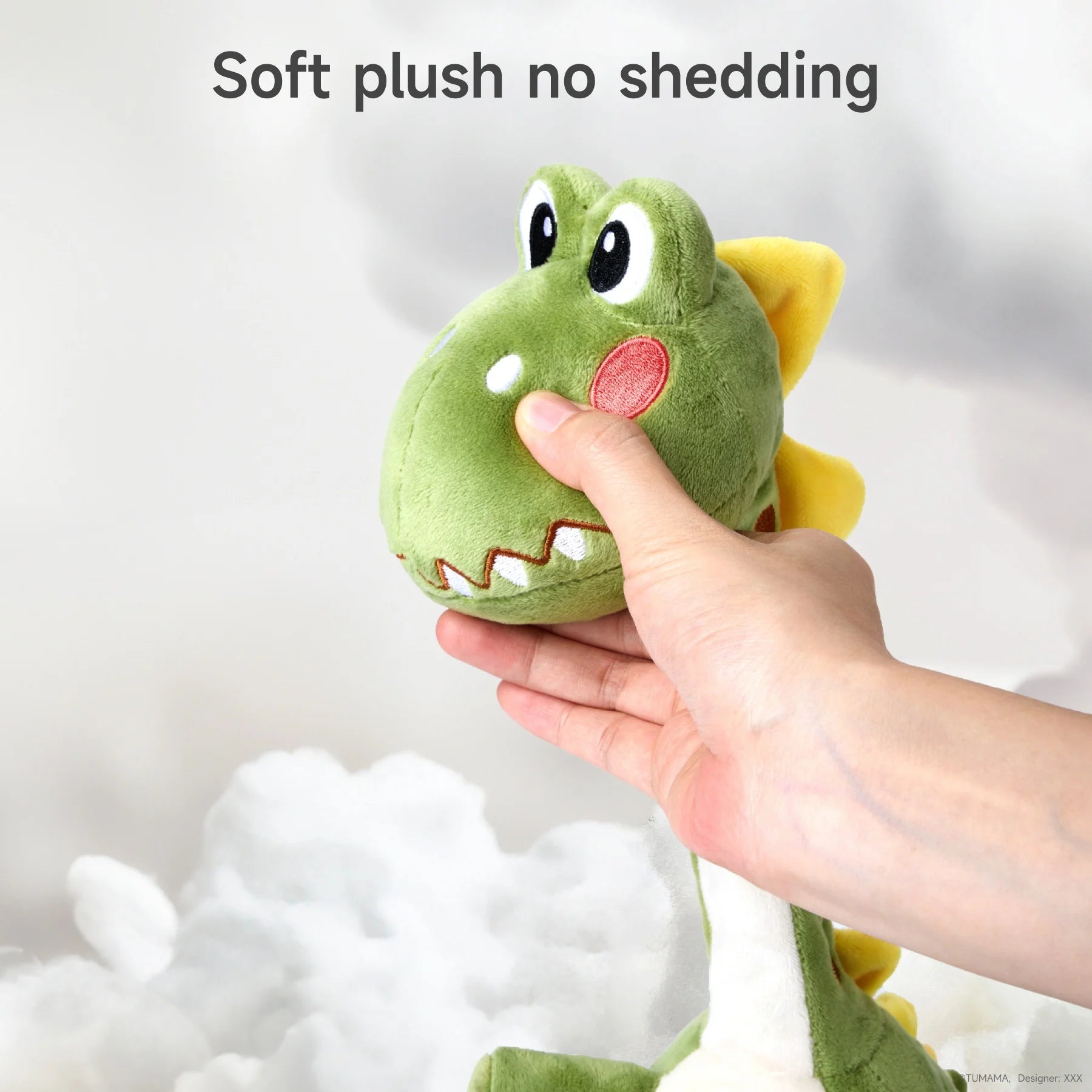 A close-up of a hand gently squeezing the head of a green plush dinosaur toy. The toy has a soft, smooth texture with vibrant details, including rosy cheeks and a happy smile, emphasizing its no-shedding and child-safe material.