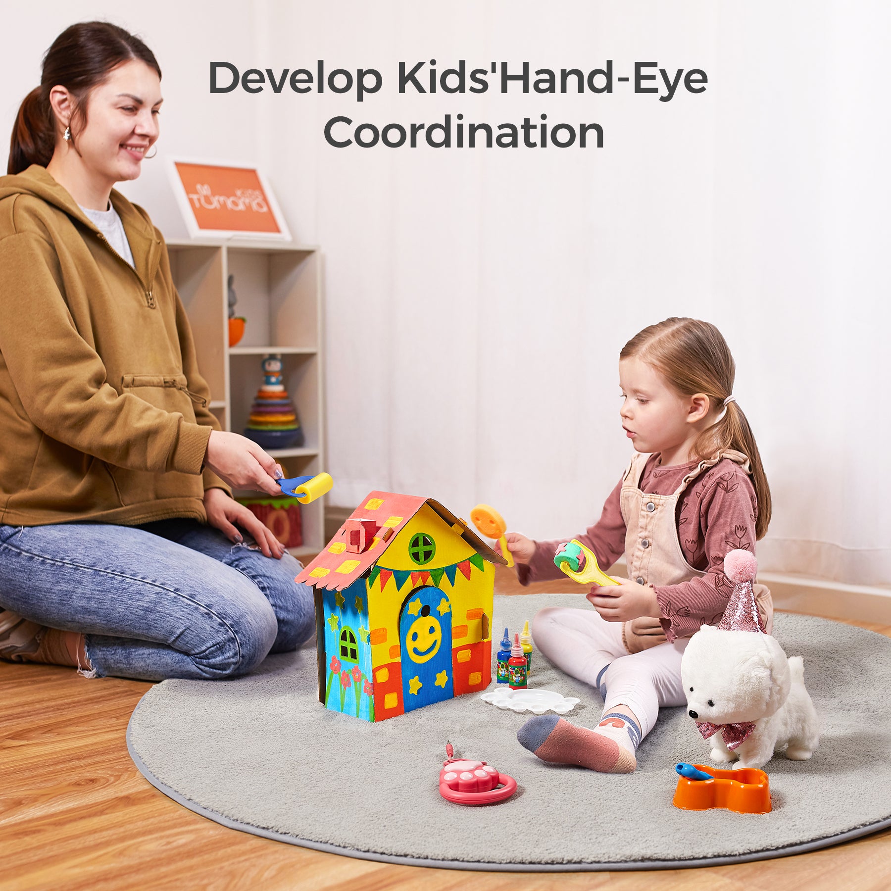 A child and an adult sitting on a rug, painting a colorful DIY cardboard playhouse. A plush puppy with a party hat and accessories is placed nearby, promoting creativity and hand-eye coordination through play.