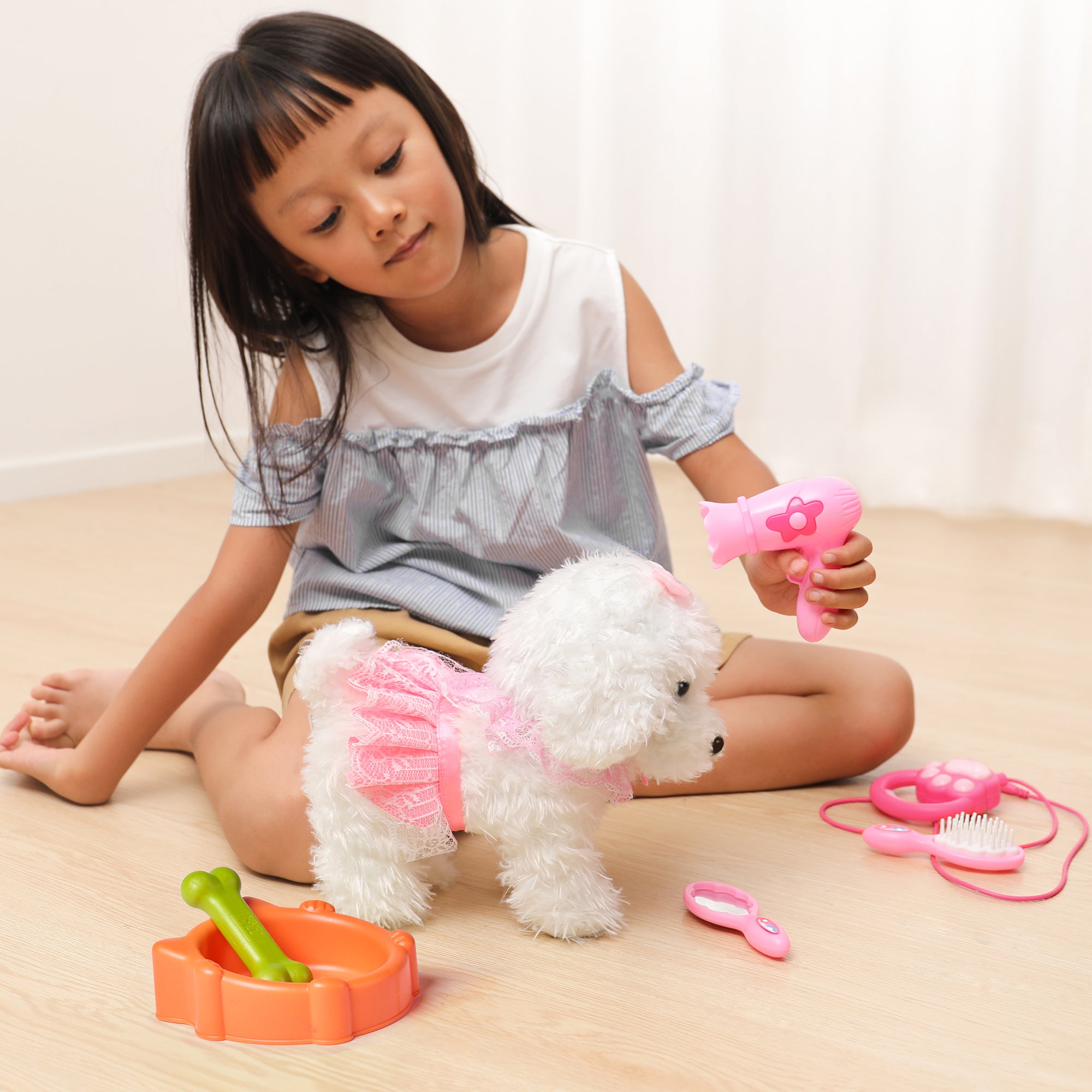 A young girl using a pink toy hairdryer to groom a white walking puppy dressed in a pink outfit. The puppy is surrounded by grooming accessories, such as a brush, hairdryer, and feeding bowl, creating an adorable and interactive playtime moment.