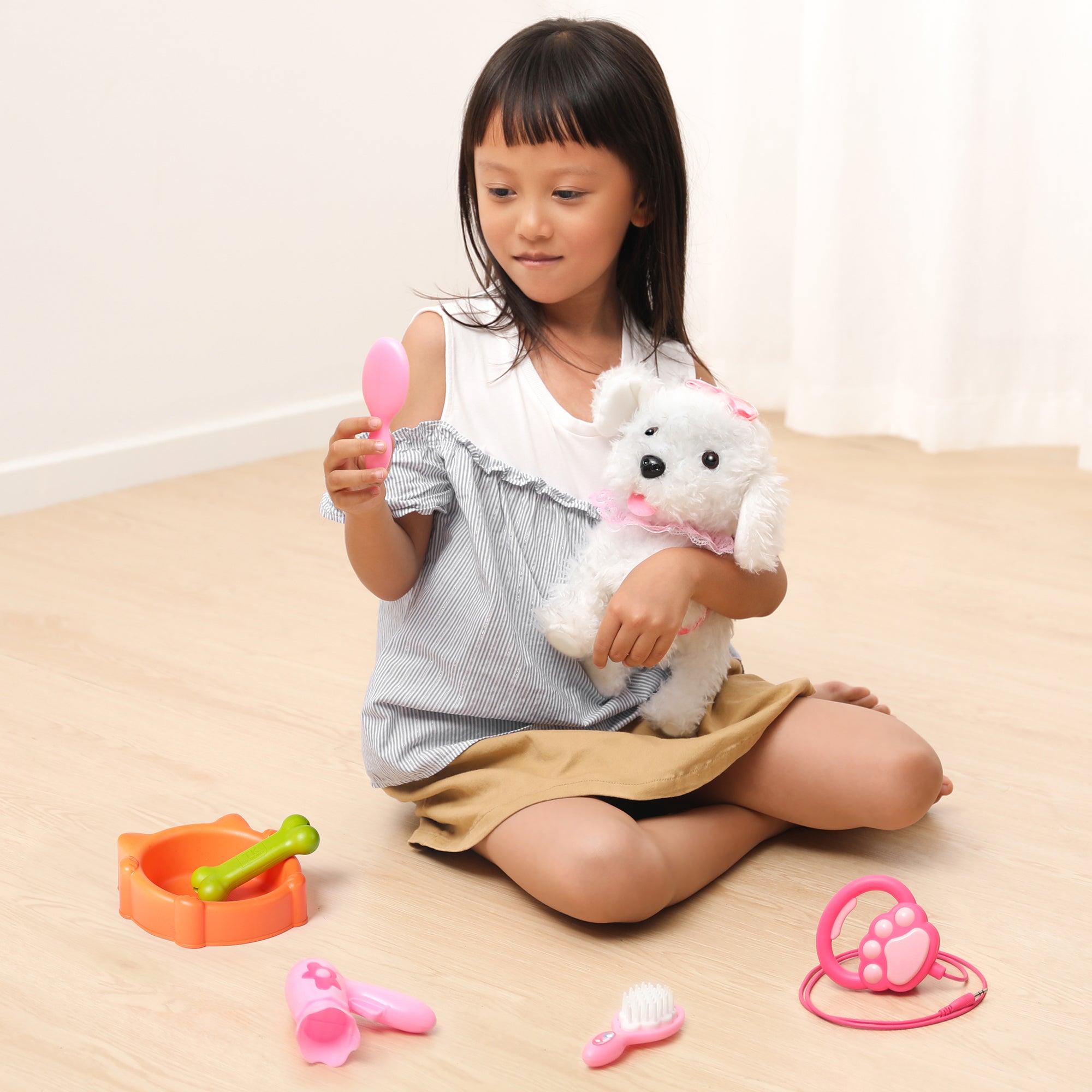 A young girl cuddling a white walking puppy toy dressed in pink, holding a pink toy mirror while surrounded by grooming accessories like a hairdryer, brush, and feeding bowl on the floor. The scene highlights imaginative and nurturing play.